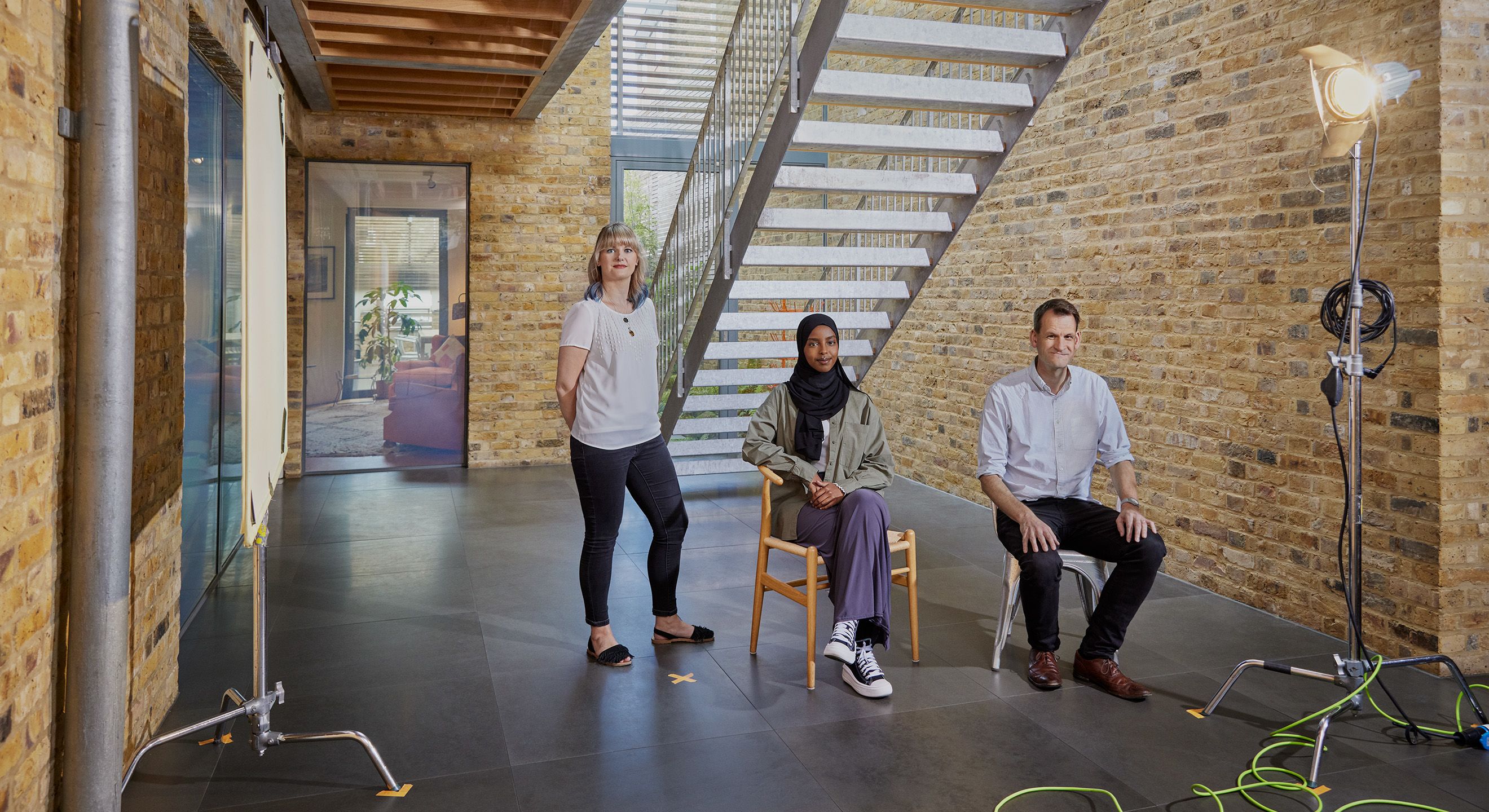 Three people sat in a studio, two sat down and one standing, all facing the camera
